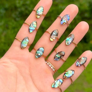 May include: A collection of eleven rings featuring marquise-shaped abalone shell inlays with iridescent green, blue, and purple hues. Ten rings have thin metal bands, while one ring features a band set with small clear stones.