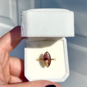 May include: A gold ring with a clear, oval-shaped centerpiece containing dried purple and white flowers. The ring is presented in an open, white velvet ring box.