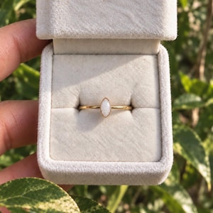 May include: A thin gold band ring featuring a single marquise-cut white opal stone set in a bezel setting. The ring is displayed inside a light-colored, velvet-lined jewelry box, showcasing the polished metal finish and the iridescent gemstone.