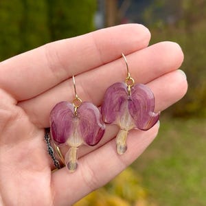 May include: A pair of handmade earrings featuring real, pressed purple flowers encased in clear resin. The earrings have gold-colored ear wires. The flower petals are a deep purple with a lighter, creamy center.