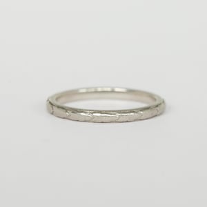 May include: A silver band ring with a textured surface. The ring has a thin profile and a continuous pattern around the band. The ring is set against a plain white background.