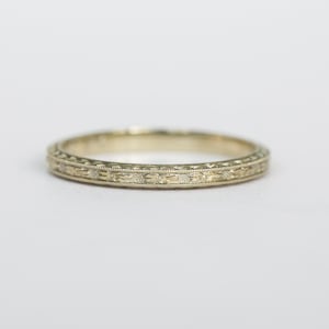 May include: A gold-colored ring with an intricate, repeating floral pattern around the band. The ring is set against a plain white background, highlighting the delicate details of the design. The ring appears to be made of metal.
