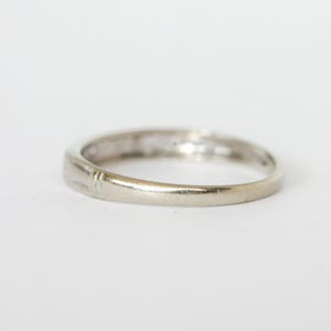 May include: A close-up of a silver ring with a smooth, polished band. The ring has a simple design with a slight curve and a few subtle markings. The ring is set against a plain white background.