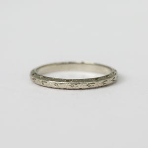 May include: A silver ring with an engraved floral pattern. The ring is thin and has a slightly worn appearance. The design is consistent around the entire band. The ring is set against a plain white background.