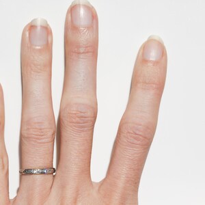 May include: Close-up of a hand with a silver ring on the ring finger. The ring has a delicate, patterned design. The hand is against a white background, with the fingers slightly spread.