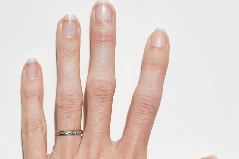 May include: Close-up of a hand with a silver ring on the ring finger. The ring has a delicate, patterned design. The hand is against a white background, with the fingers slightly spread.