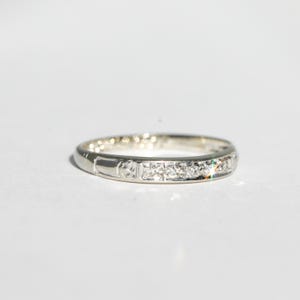 May include: A silver band ring with a series of small, clear stones set across the top. The ring has a simple, elegant design, with a smooth, polished finish. The ring is likely made of sterling silver or a similar metal.