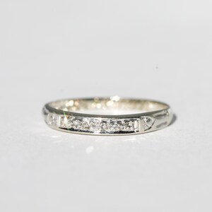 May include: A silver band ring with a series of small, clear gemstones set across the top. The ring has a delicate design with subtle patterns along the sides. The ring is shown against a white background.