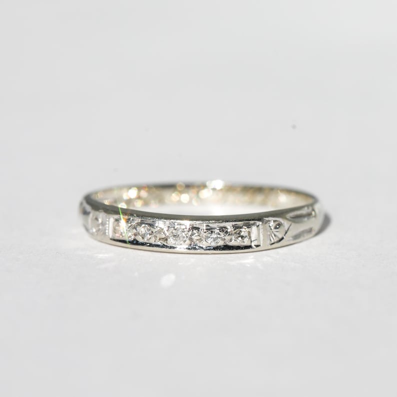 May include: A silver band ring with a series of small, clear gemstones set across the top. The ring has a delicate design with subtle patterns along the sides. The ring is shown against a white background.