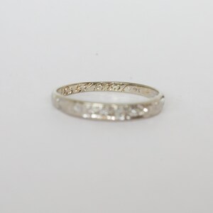 May include: A delicate, white gold ring with a repeating floral pattern and engraved text. The ring is set against a plain white background, highlighting its intricate design and craftsmanship. The ring is a classic piece of jewelry.