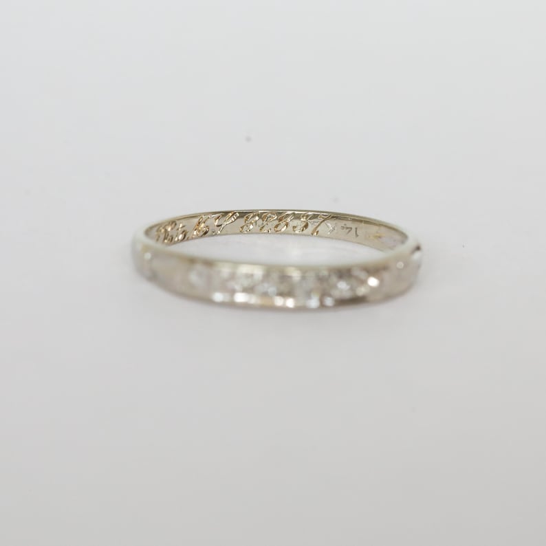 May include: A delicate, white gold ring with a repeating floral pattern and engraved text. The ring is set against a plain white background, highlighting its intricate design and craftsmanship. The ring is a classic piece of jewelry.