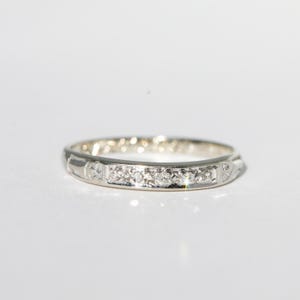 May include: A silver ring with a series of small, clear gemstones set in a row. The ring has a delicate design with subtle patterns along the band. The ring is set against a white background.