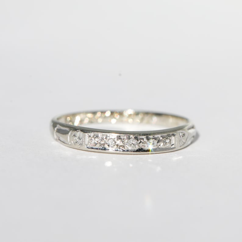 May include: A silver ring with a series of small, clear gemstones set in a row. The ring has a delicate design with subtle patterns along the band. The ring is set against a white background.