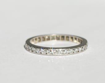 Könnte beinhalten: Ein silberfarbener Eternity-Ring mit einer durchgehenden Reihe kleiner, runder, klar facettierter Steine, die in das Metall eingefasst sind. Der Ring hat eine polierte Oberfläche und reflektiert das Licht, was die Anordnung betont.