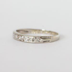 May include: A silver ring with small, clear gemstones set in a row. The ring has a simple, elegant design with a decorative pattern on one side. The ring is set against a plain white background.