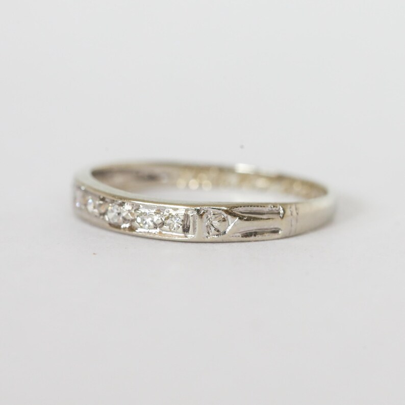 May include: A silver ring with small, clear gemstones set in a row. The ring has a simple, elegant design with a decorative pattern on one side. The ring is set against a plain white background.