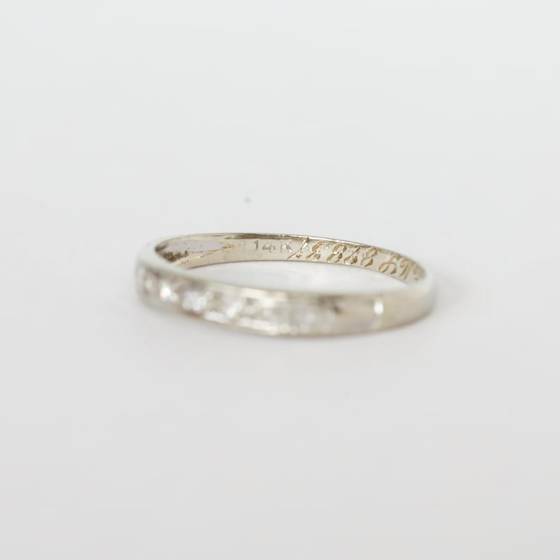 May include: A white gold ring with small, clear stones set along the band. The ring has engraved text on the inside. The ring is set against a white background.