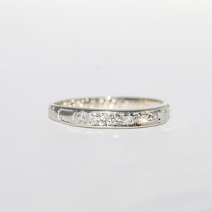 May include: A silver band ring with a row of small, clear gemstones. The ring has a simple, elegant design, with the gemstones set in a line across the top. The ring is on a white background.