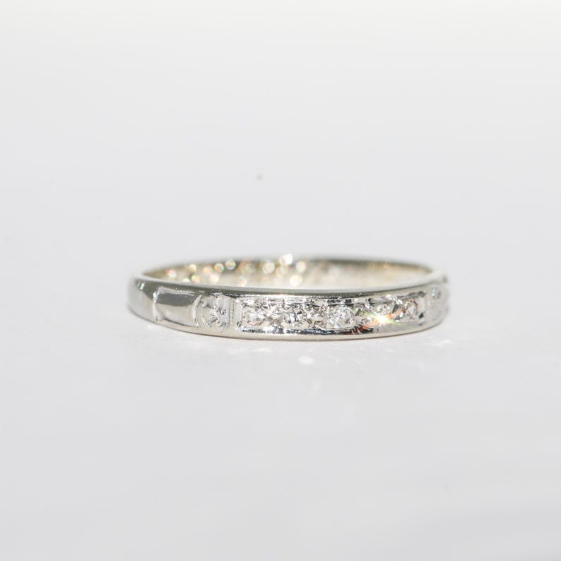 May include: A silver band ring with a row of small, clear gemstones. The ring has a simple, elegant design, with the gemstones set in a line across the top. The ring is on a white background.