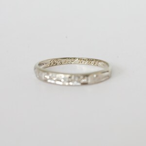 May include: A silver ring with engraved text and a decorative band. The ring is set against a white background, highlighting its intricate design and delicate details. The text on the ring is small and appears to be a date or inscription.