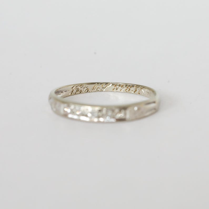 May include: A silver ring with engraved text and a decorative band. The ring is set against a white background, highlighting its intricate design and delicate details. The text on the ring is small and appears to be a date or inscription.