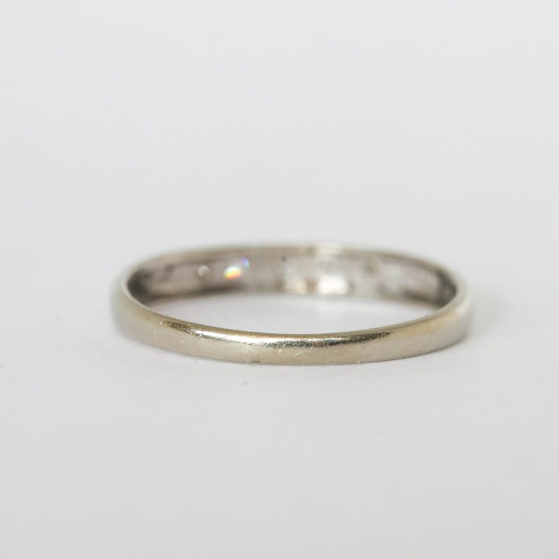 May include: A close-up shot of a silver ring with small, clear stones set into the band. The ring is set against a white background, highlighting its simple, elegant design. The ring appears to be made of a light-colored metal.