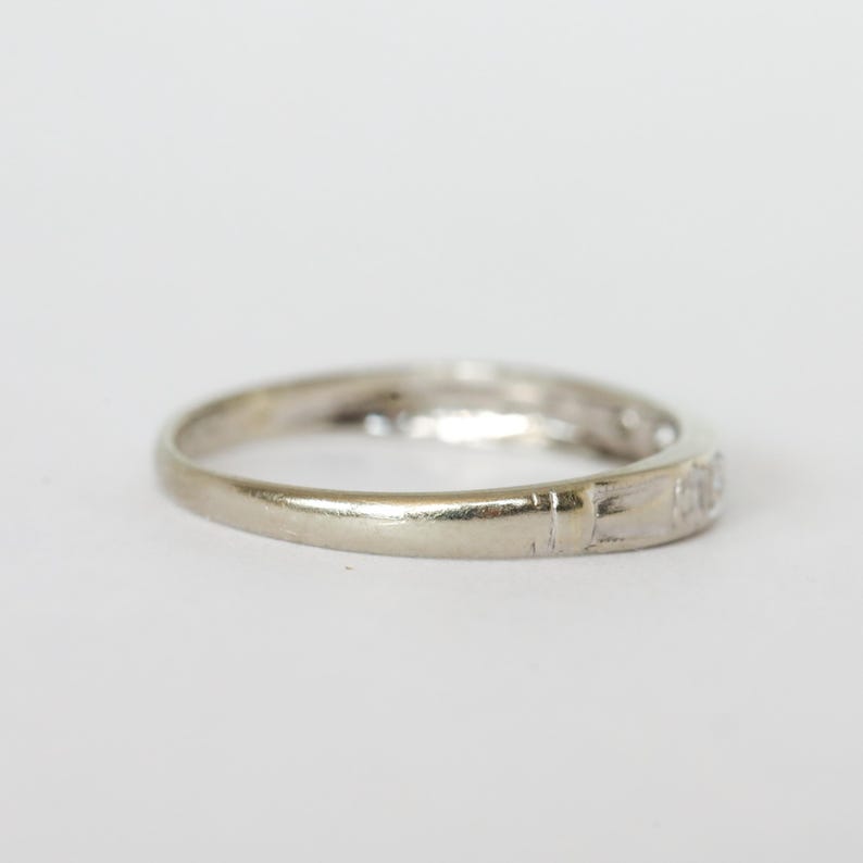 May include: A close-up of a white gold ring with small, clear gemstones set along the band. The ring is slightly worn, with visible scratches on the surface. The ring is set against a plain white background.
