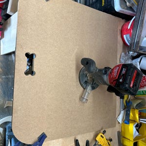 May include: A close-up of a light brown wooden board with a rounded edge and a bone-shaped cutout. A red and black electric router is attached to the board, with a clear plastic guard around the router bit. The board is secured to a workbench with blue clamps.