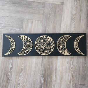 Puede incluir: Una decoración de pared de madera negra con cinco lunas de metal dorado, cada una con un diseño floral. Las lunas están dispuestas en una fila, con la luna más grande en el centro.
