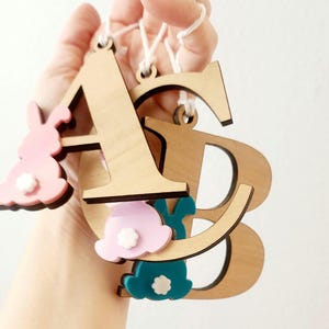 May include: Wooden letters A and B with pink and teal bunny cutouts hanging from white string. The letters are unfinished and have a natural wood color.