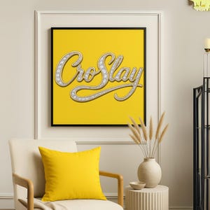 Op de afbeelding: Een ingelijste kunstdruk met de tekst "Cro Slay" in een cursief lettertype, versierd met een diamantachtig patroon, tegen een heldergele achtergrond. De print zit in een zwarte lijst en een geel kussen ligt op een stoel.