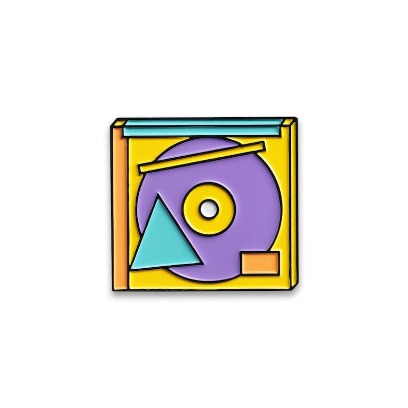 Puede incluir: Un pin de esmalte amarillo, naranja y morado con forma de casete con un disco en su interior. El disco tiene un fondo morado con un c&iacute;rculo amarillo en el centro y un tri&aacute;ngulo turquesa.