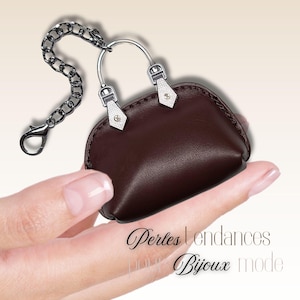 Puede incluir: Un pequeño llavero con forma de bolso marrón oscuro con detalles metálicos plateados, que incluyen una cadena y un cierre. El bolso tiene un asa curva y tiradores de cremallera decorativos. El texto "Perles tendances pour Bijoux mode" está incluido.