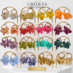 Può includere: Una collezione di sedici paia di orecchini a cerchio esposti su un supporto bianco a gradini. Ogni paio presenta cerchi color oro decorati con piccoli ciondoli a forma di fiore a petali multipli in vari colori, tra cui bianco, giallo, arancione, rosa, viola e blu.