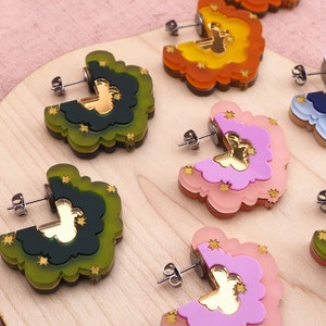 May include: A collection of colorful cloud-shaped stud earrings. Each earring features layered acrylic in shades of green, orange, pink, and blue, with gold star accents and a gold mirrored center. The earrings are displayed on a wooden surface.