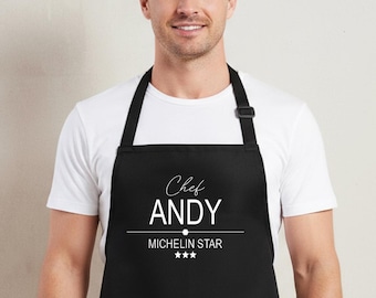 May include: A black apron with two front pockets. The apron has the text "Chef ANDY" and "MICHELIN STAR" printed in white. The apron is worn over a white t-shirt.