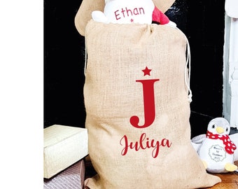 May include: A large burlap Christmas gift bag with a red initial "J" and the name "Juliya" printed on it. A Santa Claus plush toy is visible inside the bag, and a penguin plush toy is next to the bag. Two burlap stockings hang in the background.