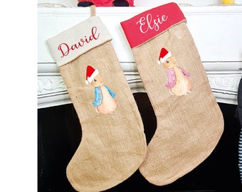 May include: Two burlap Christmas stockings featuring a printed rabbit character wearing a Santa hat. One stocking has a cream cuff with the name David, and the other has a red cuff with the name Elsie. A small elf doll with the name Giovanni is nearby.