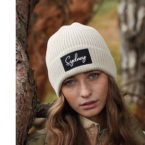 Könnte beinhalten: Eine weiße Strickmütze mit einem schwarzen Patch, auf dem "Sydney" in weißer Kursivschrift steht.