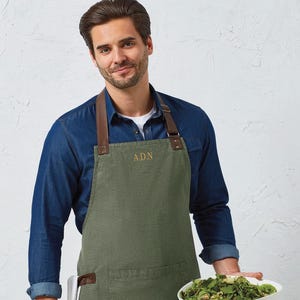 Op de afbeelding: Olijfgroene schort met bruine leren bandjes en de initialen "A.D.N." geborduurd op de borst. De schort heeft een grote voorzak. Een persoon draagt het schort en houdt een witte saladeschaal vast.
