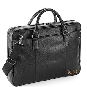 May include: Black leather briefcase with a top handle and a shoulder strap. The briefcase has a zipper closure and a gold monogram that reads "K.D."