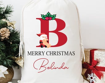 Pode incluir: Uma sacola de presente de Natal branca com fecho de cordão. A sacola apresenta uma grande letra "B" vermelha com azevinho e uma ilustração do Papai Noel. As palavras "MERRY CHRISTMAS" e o nome "Belinda" são impressas em vermelho.