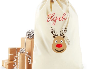 Puede incluir: Una bolsa de regalo crema con cordón, con el nombre "Elijah" en escritura roja y una cara de reno de dibujos animados. Varios regalos envueltos con lazos plateados sugieren un tema navideño.