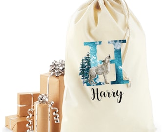 Pode incluir: Saco de presente creme com cordão e design em aquarela. O design apresenta um lobo uivando, uma letra "H" azul com flocos de neve e o nome "Harry" em escrita preta. Presentes embrulhados em papel castanho estão no fundo.