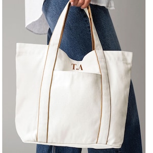 May include: A cream canvas tote bag with brown trim and handles. The bag has a front pocket and the initials "T.A" embroidered in brown. The bag is held by a person wearing blue denim jeans.