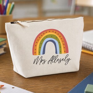 Puede incluir: Un estuche de lona color crema con cremallera que presenta un gráfico impreso de un arcoíris multicolor con arcos rojos, naranjas, amarillos, verdes y azules. El texto Mrs Allesely está impreso en cursiva negra bajo el diseño del arcoíris.