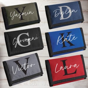 Puede incluir: Una colección de carteras personalizadas en varios colores, incluyendo verde oliva, azul marino, negro, azul, gris y rojo. Cada cartera presenta una inicial grande y un nombre en un color contrastante, con un borde negro.