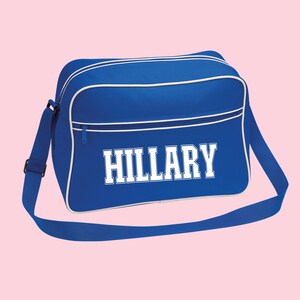 May include: A royal blue messenger bag with white trim and the name "HILLARY" in bold, white, block letters. The bag has a zippered top, a long adjustable shoulder strap, and a rectangular shape.