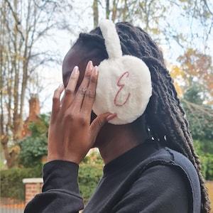 May include: White faux fur earmuffs with a pink glitter letter "E" embroidered on the side.