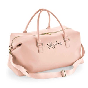 May include: A blush pink leather travel bag with a gold zipper and a black leather strap. The bag has the name "Skylar" written on the front in black cursive.
